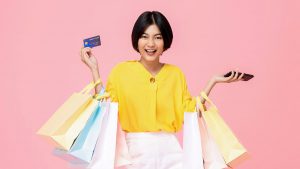 Fundo rosa. Ao centro da imagem uma mulher branca, jovem e sorridente, segurando sacolas de compra e um cartão de crédito.