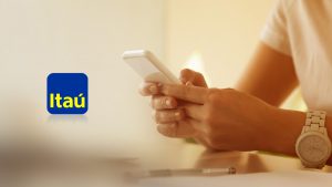 Na imagem está o logotipo do Banco Itaú e, ao lado direito, a mão de uma mulher mexendo em um celular, como se estivesse fazendo uma transferência online.