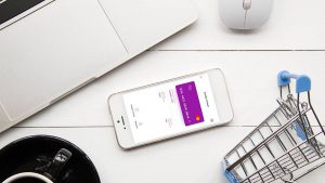 Foto de uma mesa contendo um notebook, uma xícara de café, um mini carrinho de compras e um celular. Na tela do aparelho está o App Nubank, com o cartão virtual.