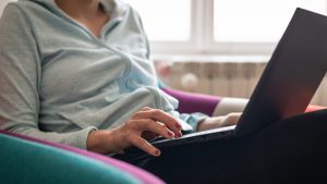 Imagem de uma mulher sentada em uma poltrona, com um moletom azul, mexendo em um notebook.