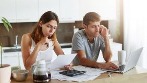 Imagem de um casal composto por um homem e uma mulher, sentados em uma mesa. O homem está olhando para um notebook e a mulher, segurando um documento, que representa a segunda via RGE.