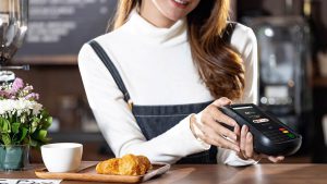 Mulher em um lugar que aparenta ser um restaurante, com um salgado em sua frente e realizando o pagamento da conta com a maquininha de cartão de crédito da C6 Bank.