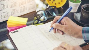 Mão masculina segurando uma caneta azul e escrevendo em um caderno.