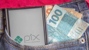 Foto de um bolso de uma calça jeans, cujo interior tem um celular e notas de R$ 100 e R$ 50. Na tela do celular está aberto o aplicativo do Pix.