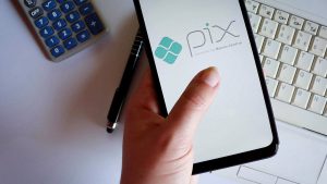 Mão feminina segurando um celular, cuja tela apresenta o aplicativo do Pix.