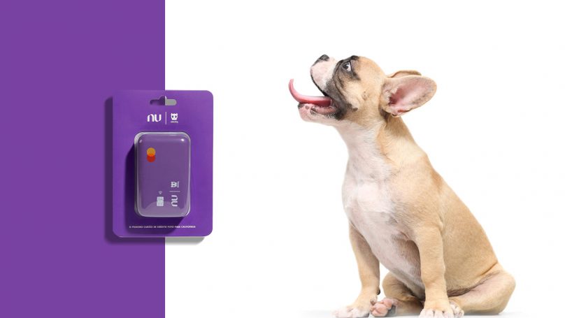 Foto de um cachorro da raça buldogue francês, olhando para o lado e com a língua para fora. Do lado esquerdo da imagem o Nudog, brinquedo em formato de cartão de crédito do Nubank.