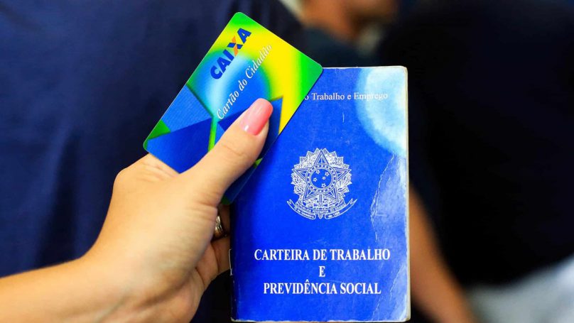 Foto da mão de uma mulher segurando uma carteira de trabalho e o cartão Caixa Cidadão para saque do Pis Pasep.