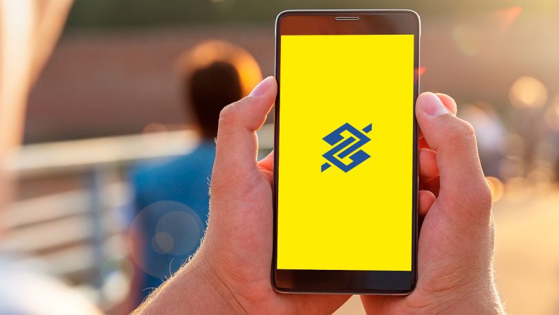Imagem de uma pessoa segurando um celular, cuja tela mostra o logotipo do Banco do Brasil.