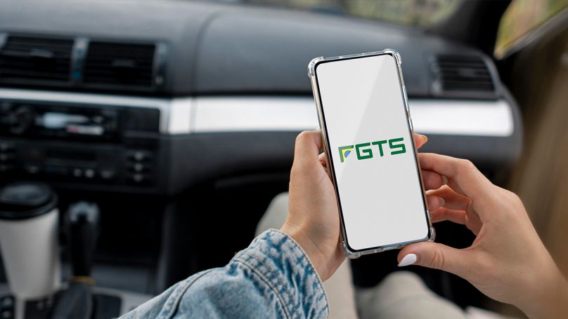 Mulher dentro de um carro, segurando um telefone celular, cuja tela apresenta o logotipo do FGTS.