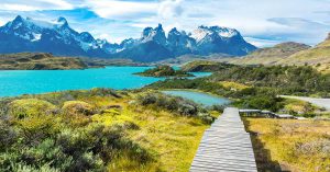 Paisagem do Chile com montanhas e áreas verdes, perto de um lago azul e de uma ponte pequena de madeira.