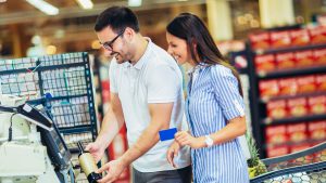 Imagem com muitas prateleiras ao fundo em desfoque, representando um mercado, com um homem e mulher passando as compras e a mulher com o cartão de crédito na mão.