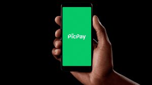 Foco na mão de uma pessoa segurando um celular, cuja tela apresenta o logotipo do PicPay