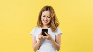 Imagem de uma mulher de camiseta branca, mexendo em um celular, como se estivesse abrindo a sua conta universitária do Banco do Brasil.