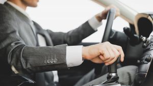 Homem de terno dirigindo um carro, com uma das mãos no volante e a outra no câmbio CVT.