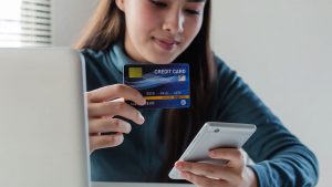 Mulher com um notebook ao lado, segurando um celular com uma mão e um cartão de crédito com a outra.