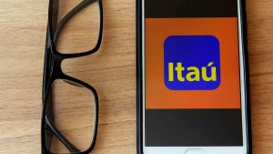 Na imagem está um óculos de grau sob uma mesa e, ao lado direito, um celular, cuja tela mostra o logotipo do Banco Itaú.