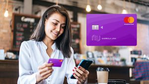 Foto de uma mulher sentada em uma cafeteria, segurando um cartão de crédito Nubank em uma mão e um celular em outra mão.