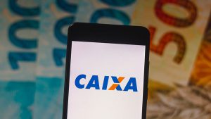 Imagem de um celular, cuja tela apresenta o logotipo da Caixa Econômica Federal. No fundo da imagem existem notas de R$ 50 e R$ 100.