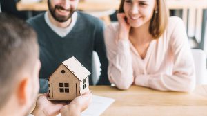 Casal sorridente conversando com um vendedor, que está segurando uma casinha de madeira com as mãos – algo que representa o financiamento imobiliário da Caixa ou Santander.