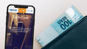 Imagem de um celular, cuja tela apresenta o site da Caixa Econômica Federal, e ao lado, uma carteira com dinheiro.