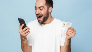 Homem sorrindo, segurando um celular em uma mão e um cartão de crédito em outra.