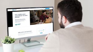 Na imagem está um homem com camisa branca, sentado de frente para um computador, como se estivesse solicitando o empréstimo do Mercado Pago, de forma online.