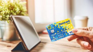 Imagina de uma mão segundo o cartão OuroCard Facil em frente a um tablet