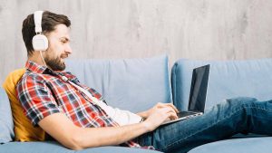 Na imagem está um homem sentado no sofá, com um fone de ouvido e mexendo em um notebook, como se estivesse na internet, procurando pelo código Nubank.