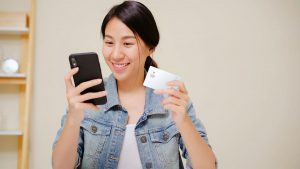 Foto de uma mulher sorridente segurando um telefone celular em uma mão e o cartão C6 Bank na outra.