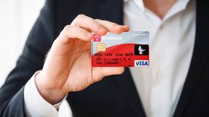Imagem de uma mão masculina segurando o cartão de crédito Bradesco. O cartão possui as cores cinza, vermelho e preto e o símbolo da bandeira Visa.