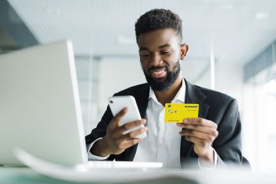 Homem negro segurando um cartão de crédito Ourocard em uma mão, e na outra um celular.