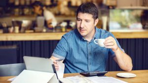 Imagem de um homem sentado, tomando um café, e mexendo em um notebook, como se estivesse consultando a 2ª via da conta Ituran pela internet.