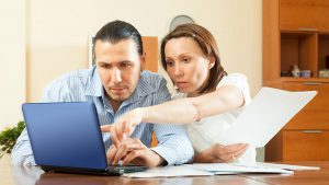 Foto de um casal composto por um homem e uma mulher, de frente para um notebook. Na imagem a mulher está com um papel na mão e apontando para o notebook, representando a negociação de dívidas na plataforma online da Casas Bahia.