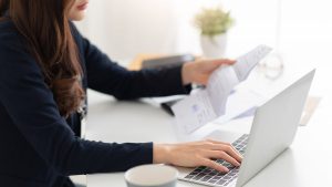 Mulher sentada de frente para um notebook com uma das mão sobre o teclado e na outra, segurando um documento que representa a segunda via da conta EDP.