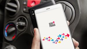 Na imagem está um homem, dentro de um carro, segurando um celular, cuja tela apresenta o aplicativo da Minha Conta Sem Parar.