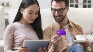 Foto de um casal sentado no sofá. Mulher usando um computador enquanto o homem segura um cartão Nubank Gold.