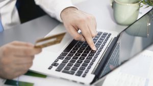 Na imagem está ilustrada uma mão feminina segurando um cartão de crédito e em um computador, como se estivesse pagando uma conta da Vivo, de forma online, com o cartão.