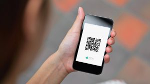 Imagem com fundo desfocado e uma mão segurando um celular com o aplicativo do Pix aberto e o QR Code na tela.
