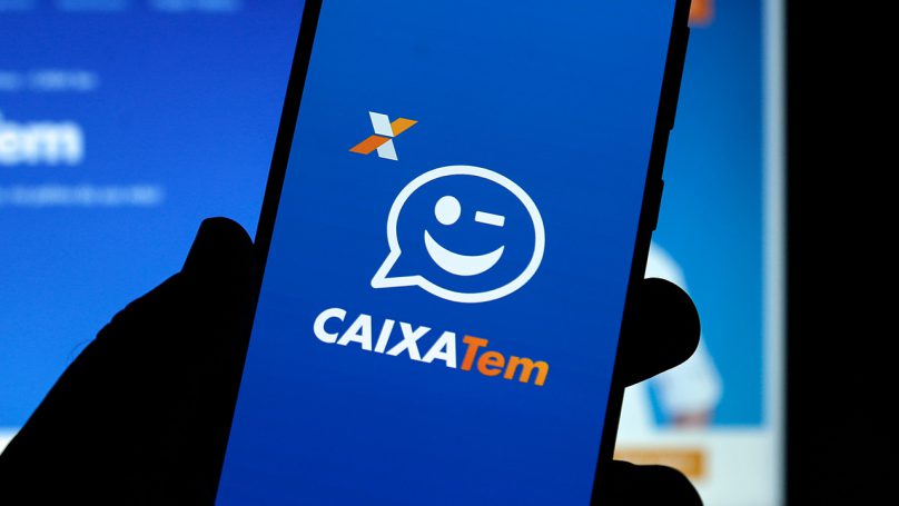 Mão segurando um telefone celular, cuja tela apresenta o logotipo da Caixa Tem.