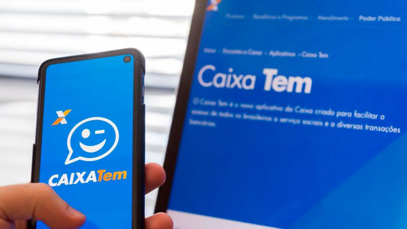 Foto da tela de um notebook conectada no site do Caixa Tem. Ao lado uma pessoa segurando um celular, na tela o Aplicativo Caixa Tem,