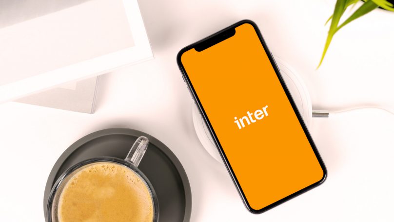 Na imagem está uma xícara de café e, ao lado direito, um celular cuja tela apresenta o logotipo do Banco Inter.
