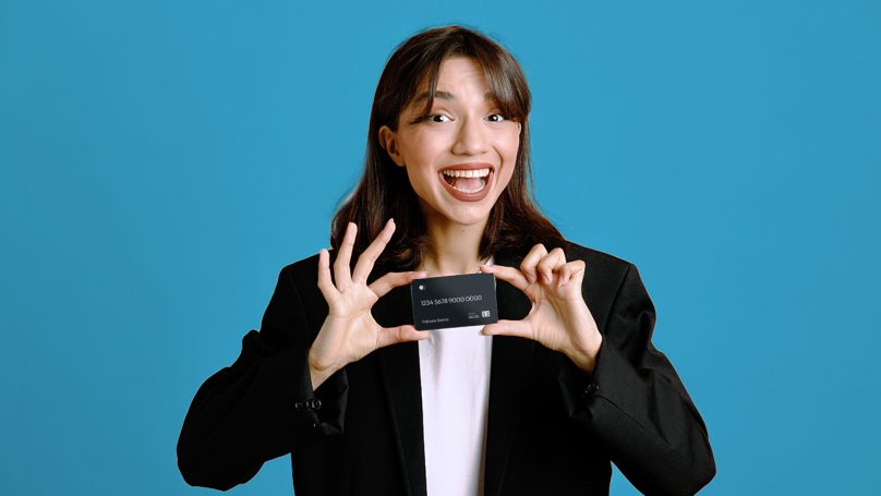 Mulher sorridente, com um sobretudo preto e uma camisa branca, segurando um cartão de crédito Platinum.